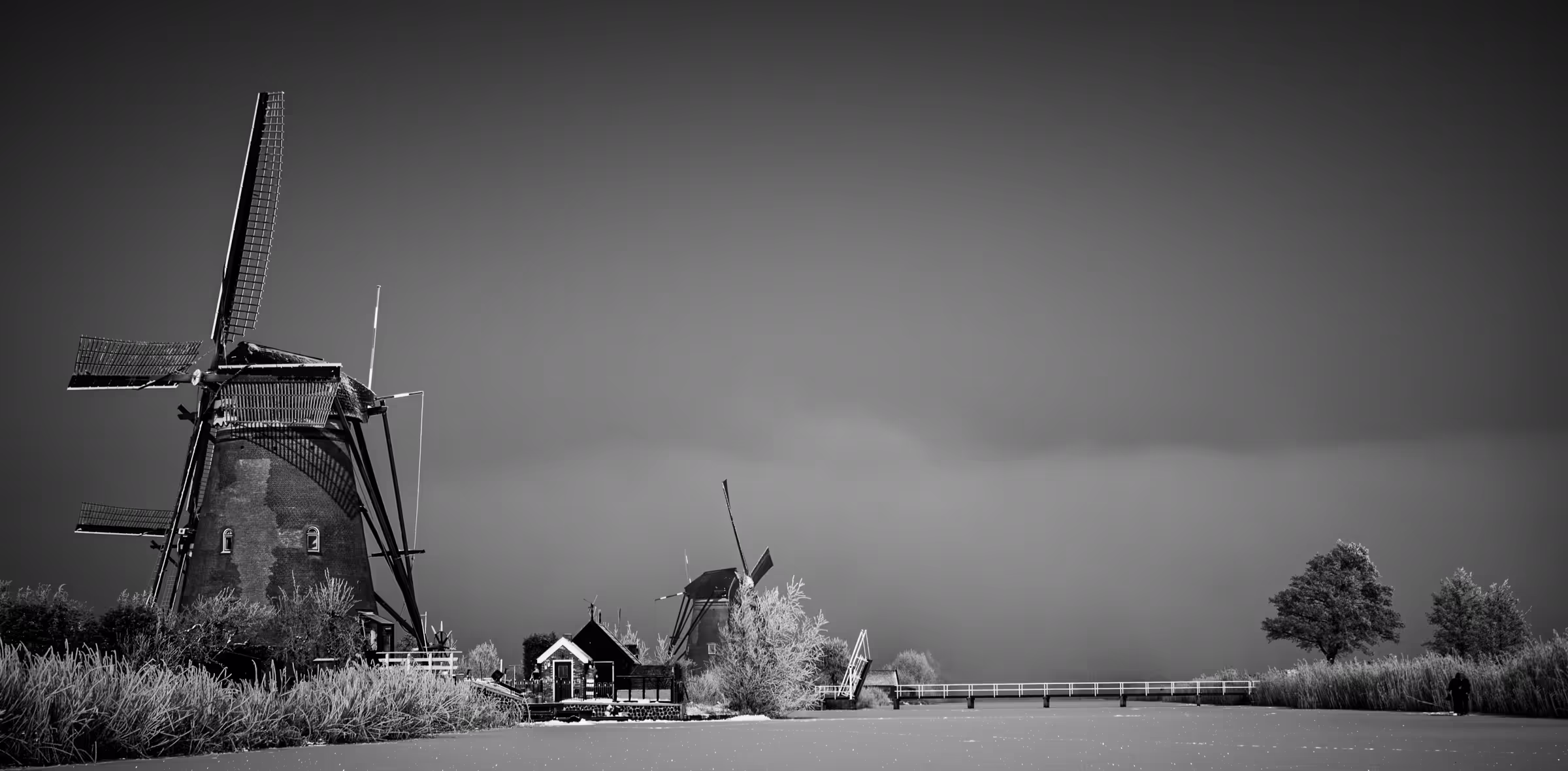 Black and white landscape photograph of windmills at Kinderdijk, Netherlands. Silhouetted windmills stand against a misty sky, with a small bridge and traditional Dutch architecture visible in the mid-ground.
